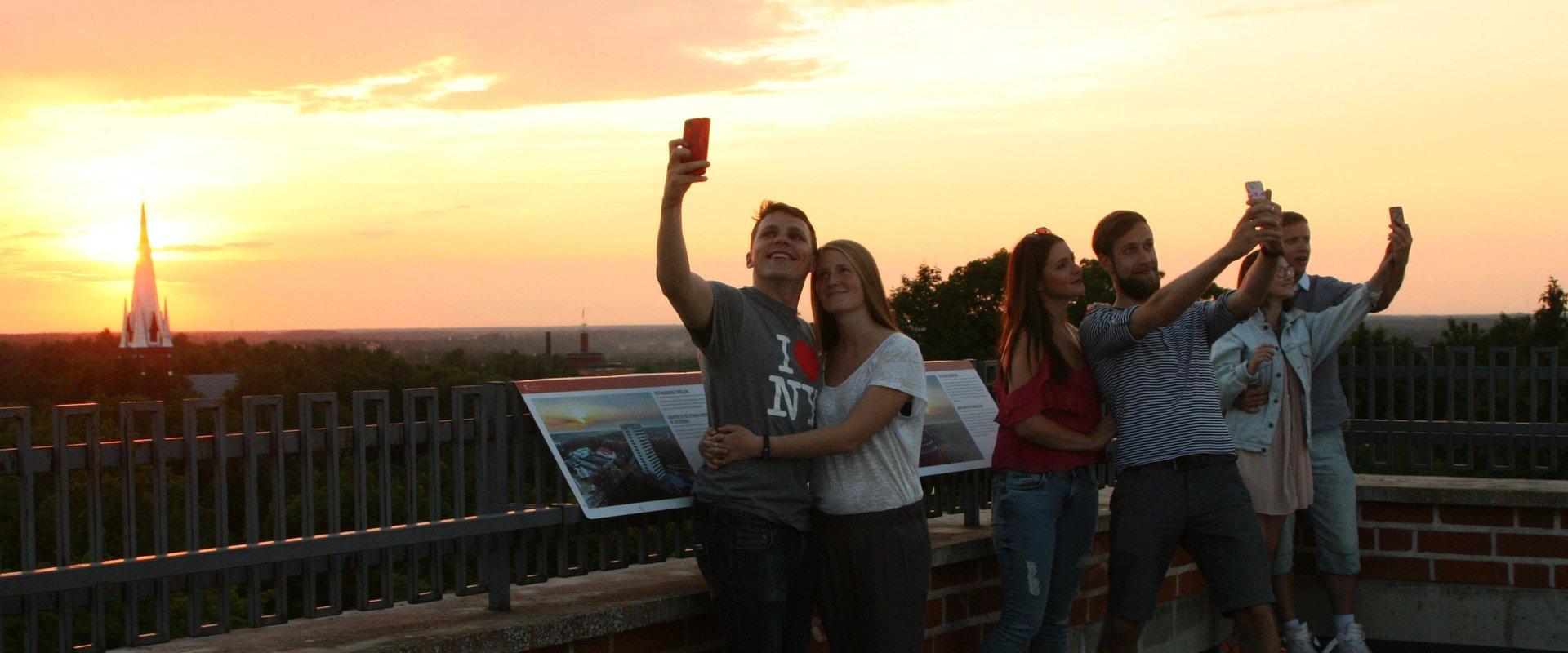 Tartu Universitātes muzejs, doma baznīcas torņi,
jaunieši saulrietā uzņem selfijus
