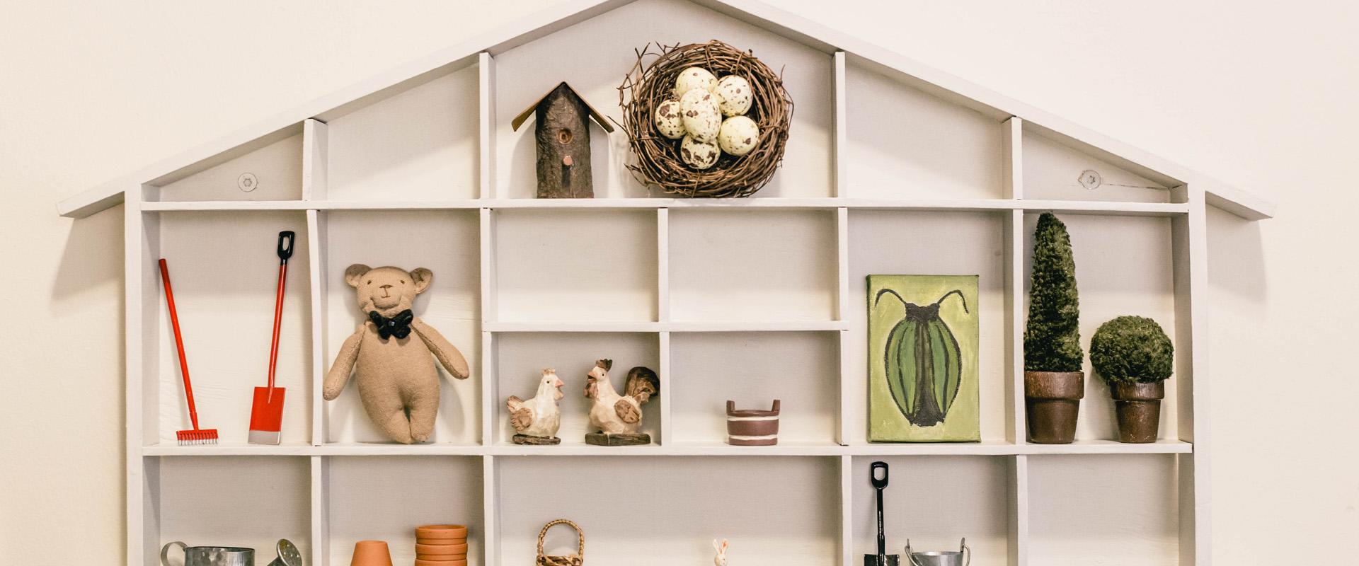 Studio shop Karud ja Pojad, tiny house-shaped shelf with miniatures, bird's nest, birdhouse, small bear, tiny rake and shovels, rooster, chicken, beetle painting, textile potted plants, bucket, watering can, flower pots