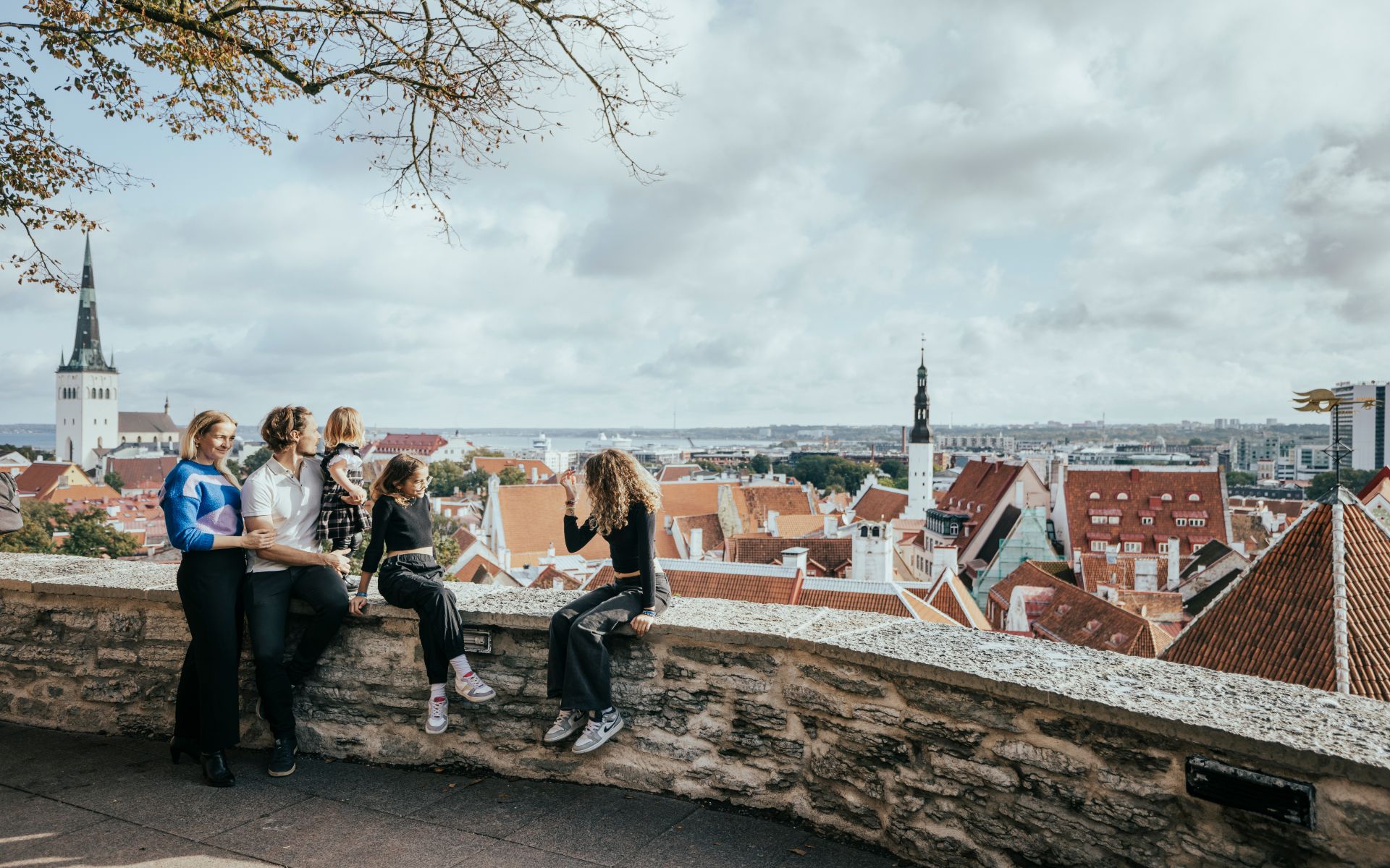 A group of four people enjoying a scenic view over Tallinn, Estonia, with medieval rooftops and the spire of a church in the background under a partly cloudy sky.