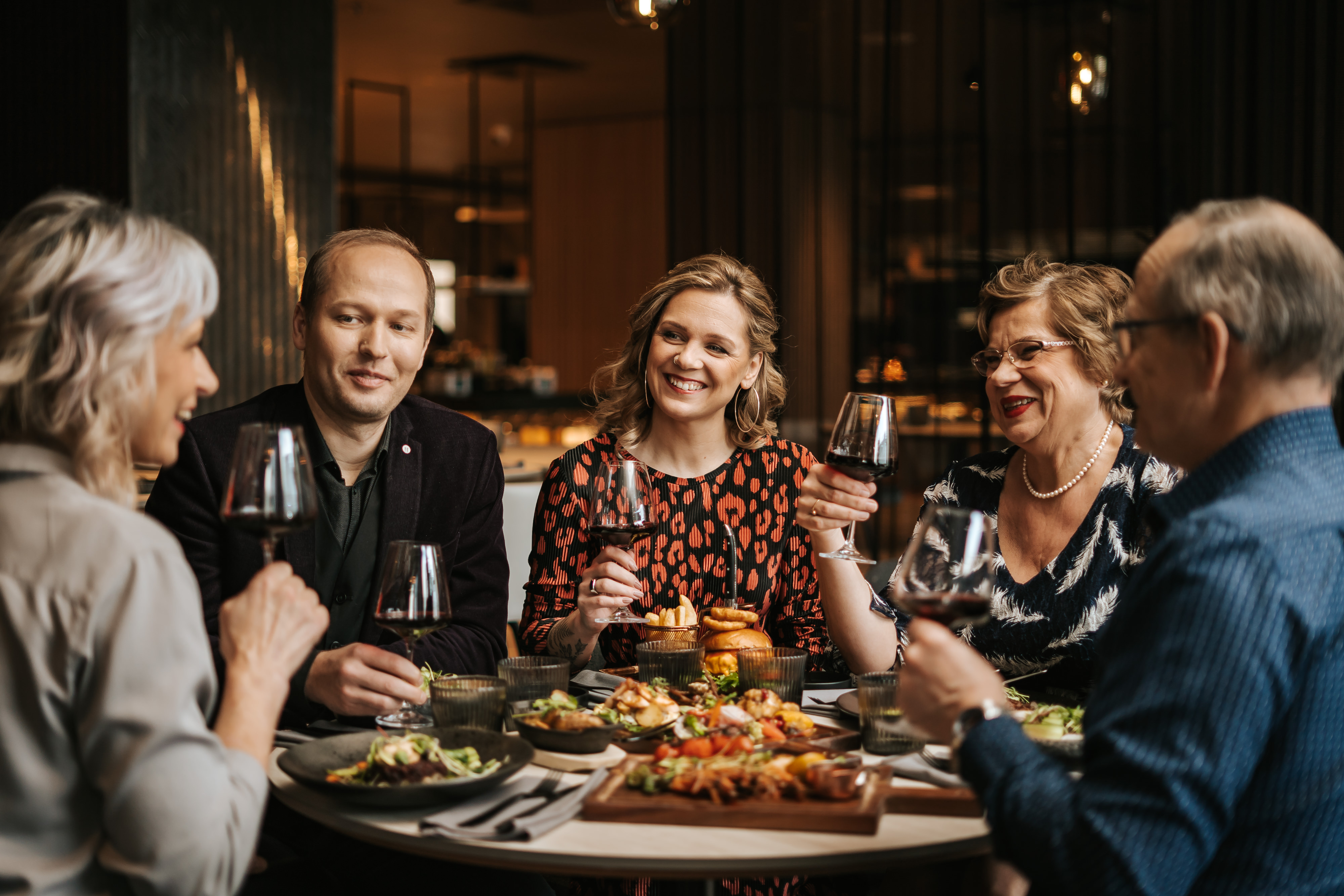 Erksates riietes viieliikmeline perekond naudib lauas koosviibimist. Käes on punaveinipokaalid.