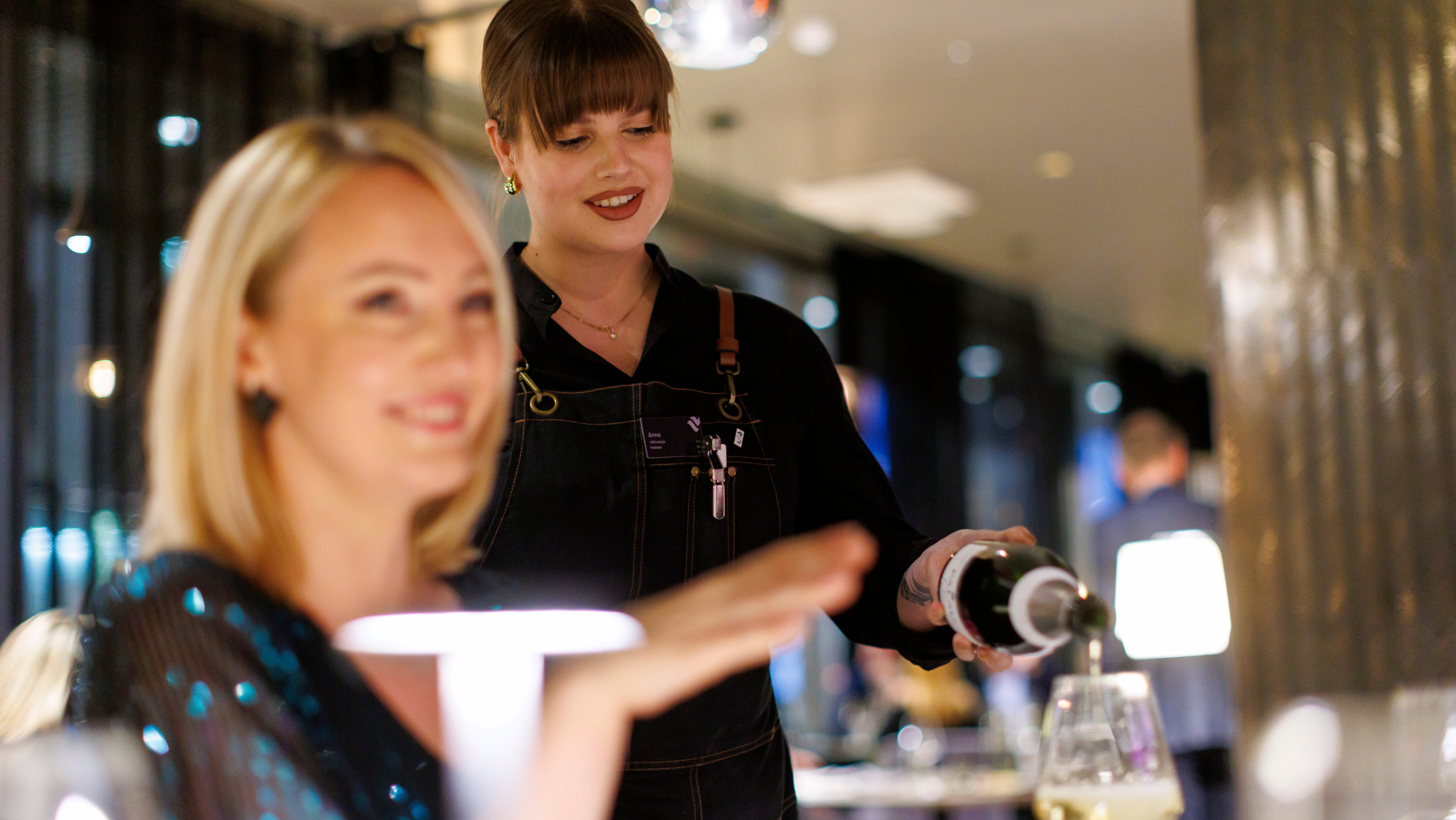 Brünett teenindaja täidab heledajuukselise kliendi pokaali vahuveiniga, mõlemad naeratavad.
