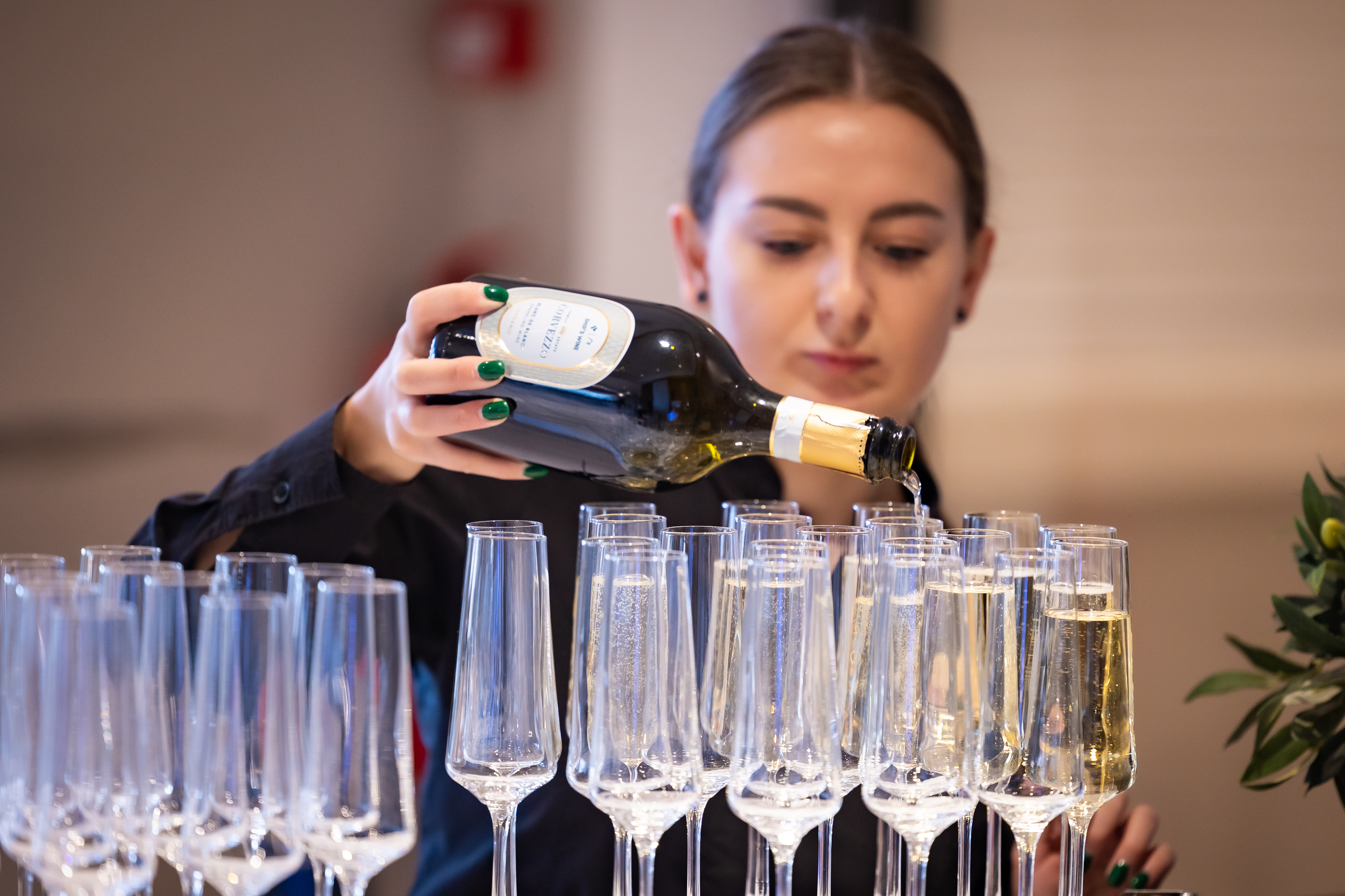 Kelner valav pokaalidesse vahuveini, fookuses on tume Cava pudel, taustal hajusalt brünett neiu.