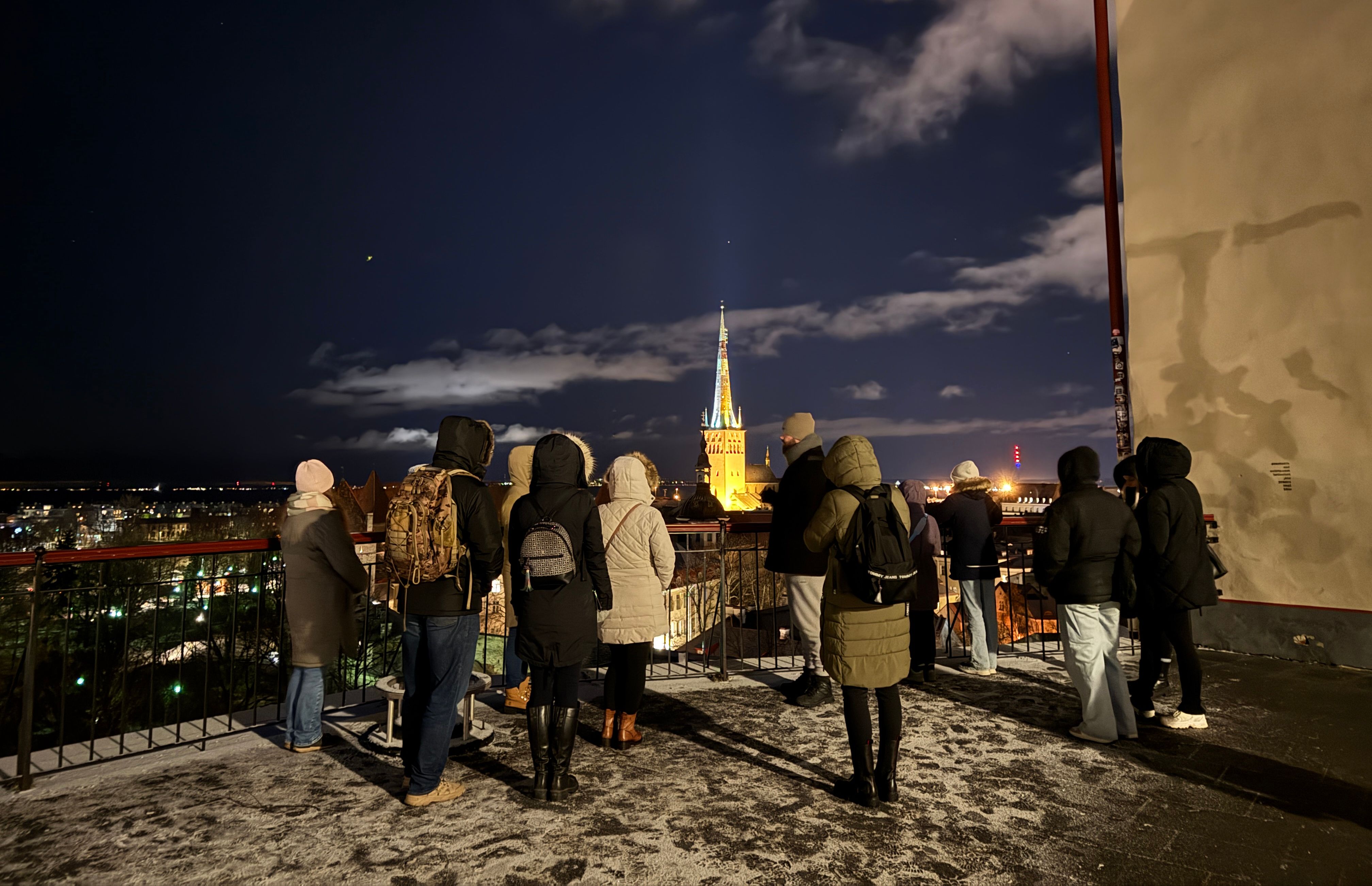 Seltskond on pimedas vanalinna vaateplatvormil taustal valgustatud Oleviste kirik