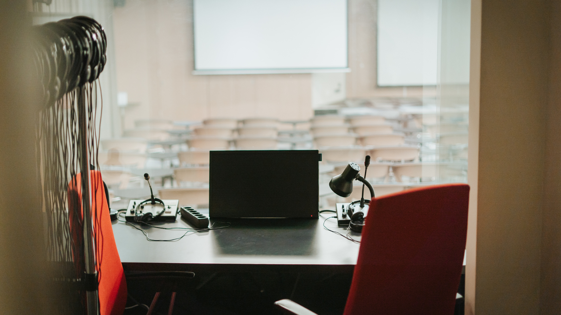 Tõlkeruum laua, tooli ja tehnikaga, taustal konverentsil osalejatele kõrvaklapid.